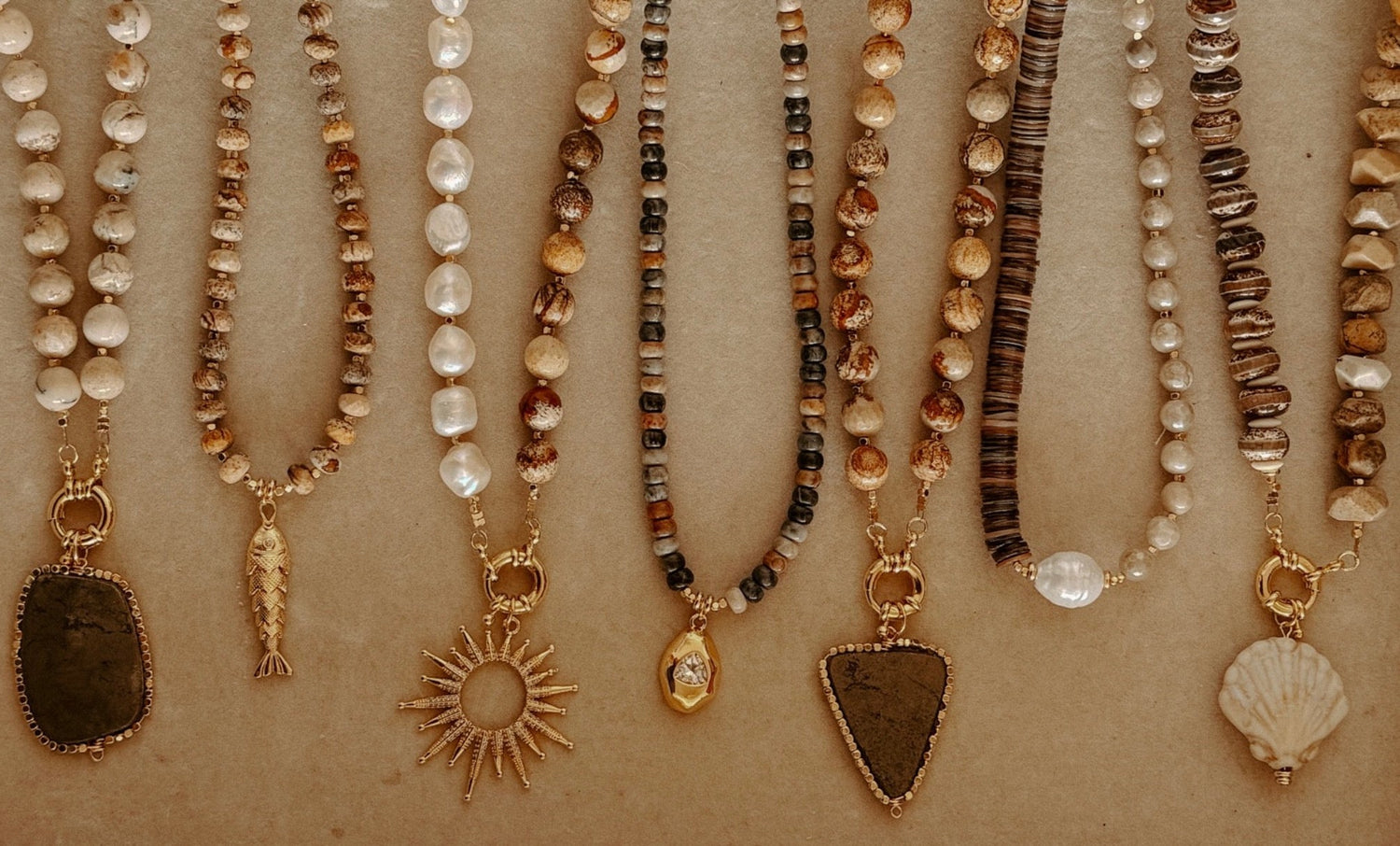 A collection of bohemian-style beaded necklaces laid out diagonally on a textured tan background. The necklaces feature various beads including white pearls, brown and beige marbled beads, striped beads, and natural stone pendants in gold-toned settings, including a shell, triangle, sun, fish, and rectangular shapes.